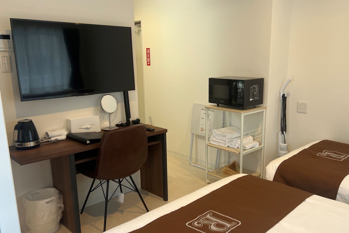 A hotel room with a desk, a television, a microwave, a bed with a brown bedspread, and a small shelf with towels.