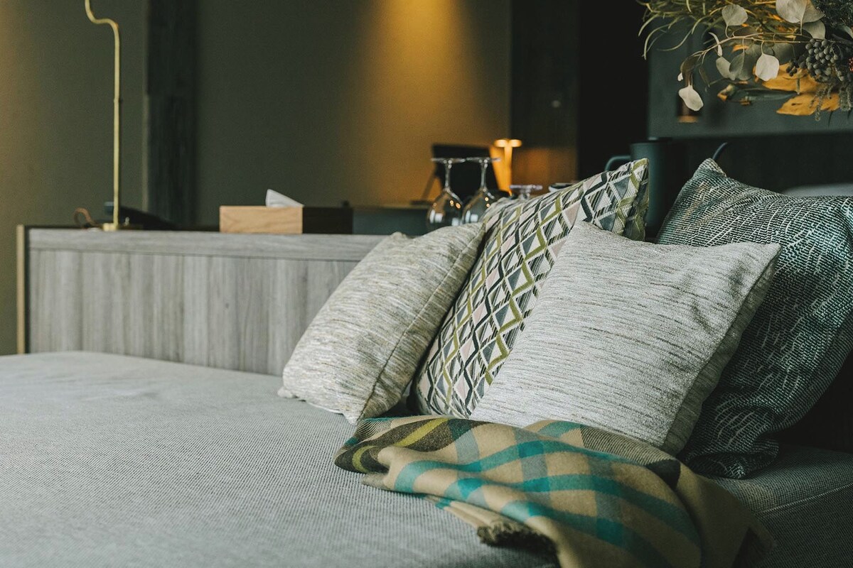 A neatly made bed with a patterned blanket and pillows, a bedside table with a lamp, and a wall-mounted shelf with decorative items.