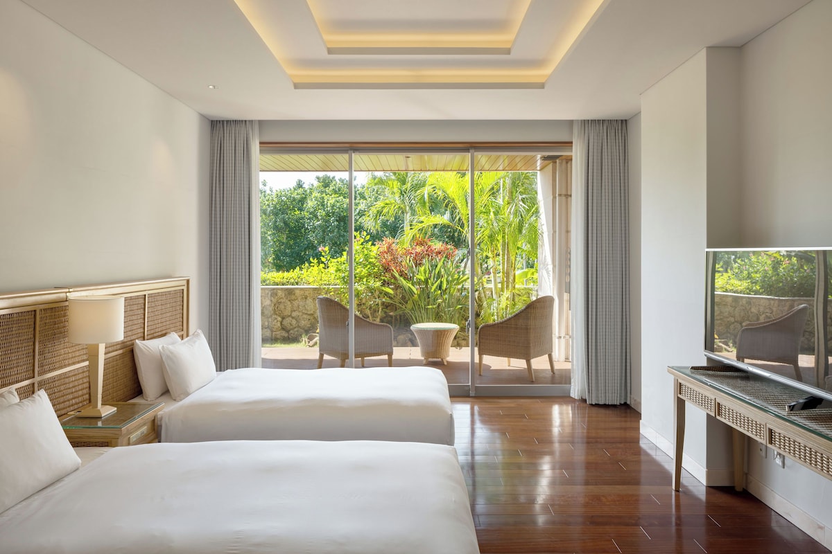 A hotel room with two beds, a TV, and a balcony with chairs and plants.