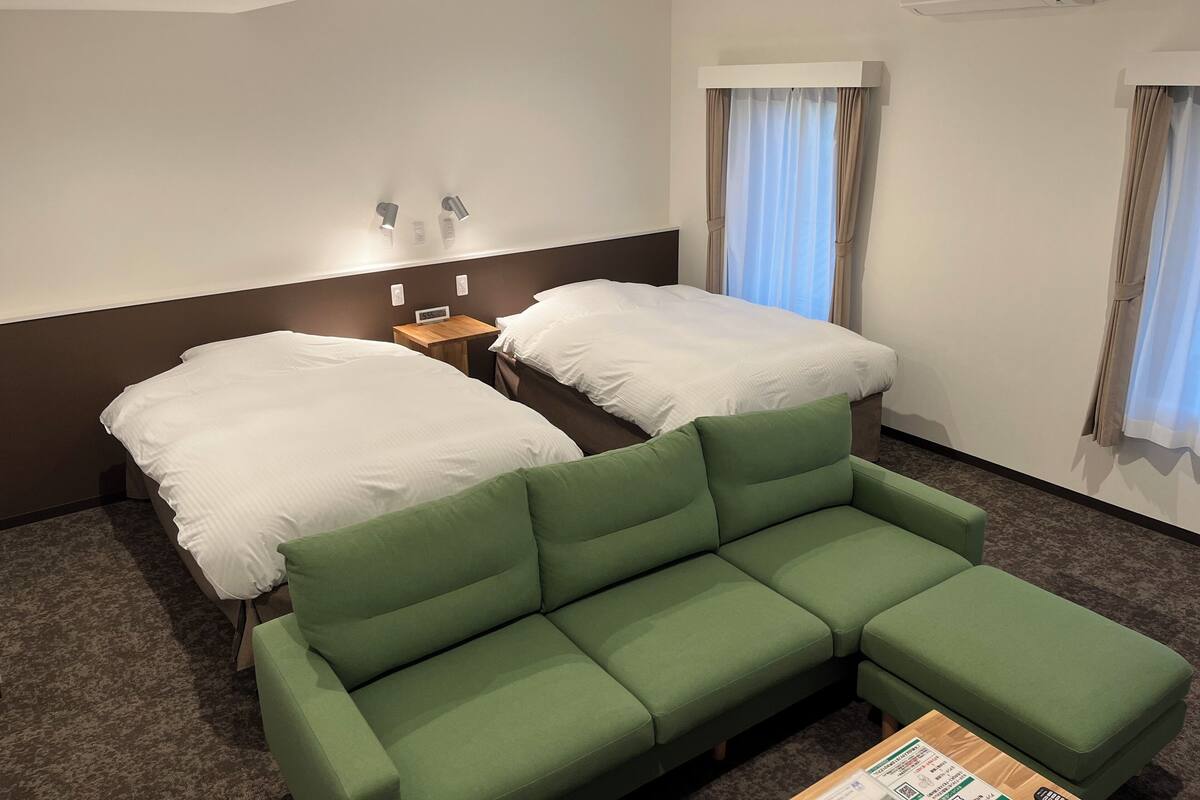 Ein Hotelzimmer mit zwei Betten, einem grünen Sofa, einem Nachttisch mit einer Uhr und einem Fenster mit Vorhängen.