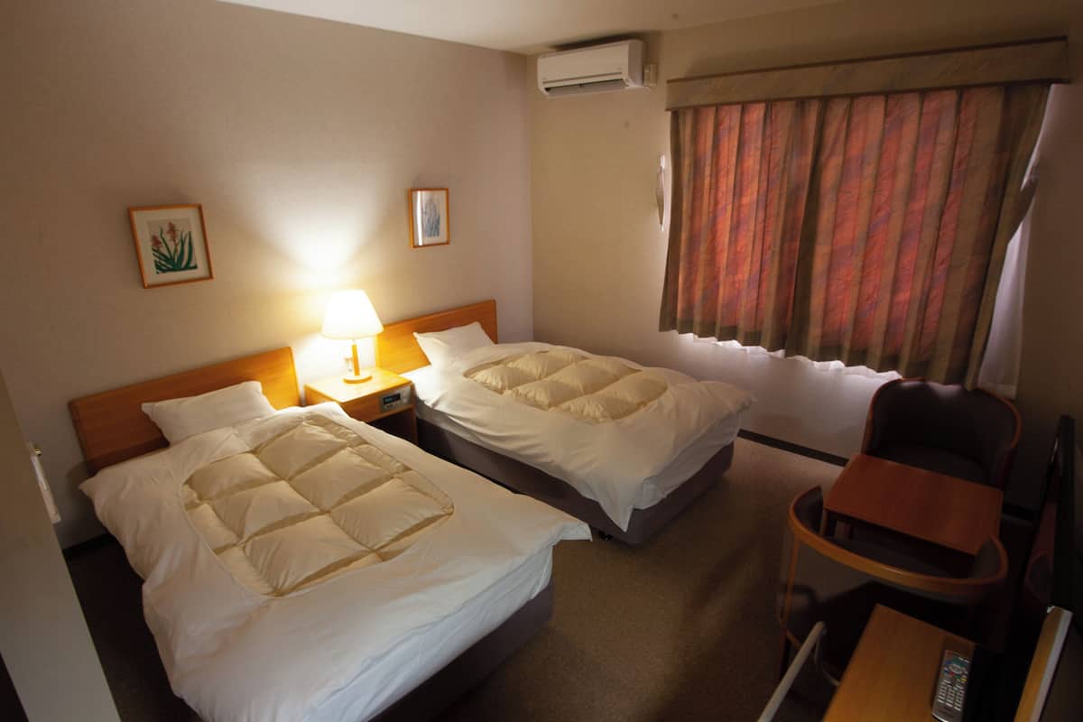 Ein Hotelzimmer mit zwei Betten, einem Nachttisch mit Lampe, einer Klimaanlage, einem Fenster mit Vorhängen und einem Sessel.