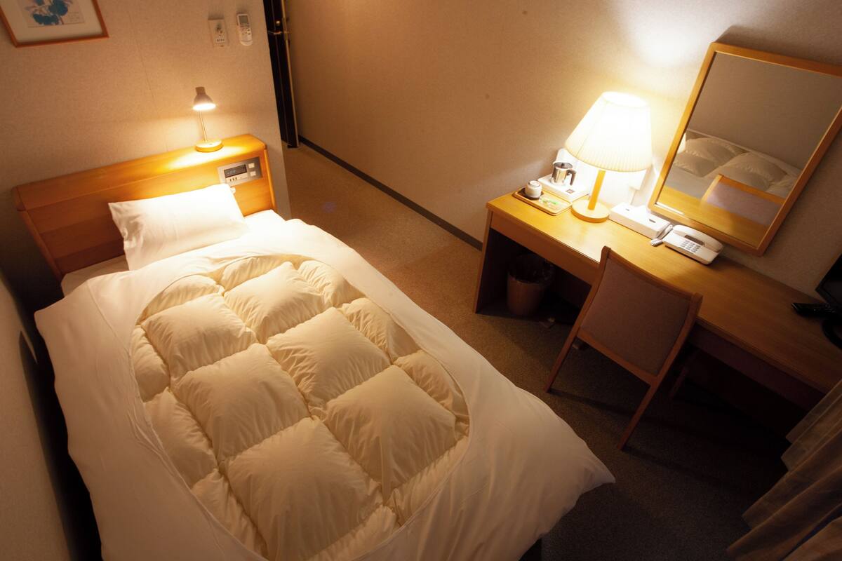 Ein Hotelzimmer mit Bett, Nachttisch mit Lampe, Stuhl, Schreibtisch mit Spiegel und Telefon.