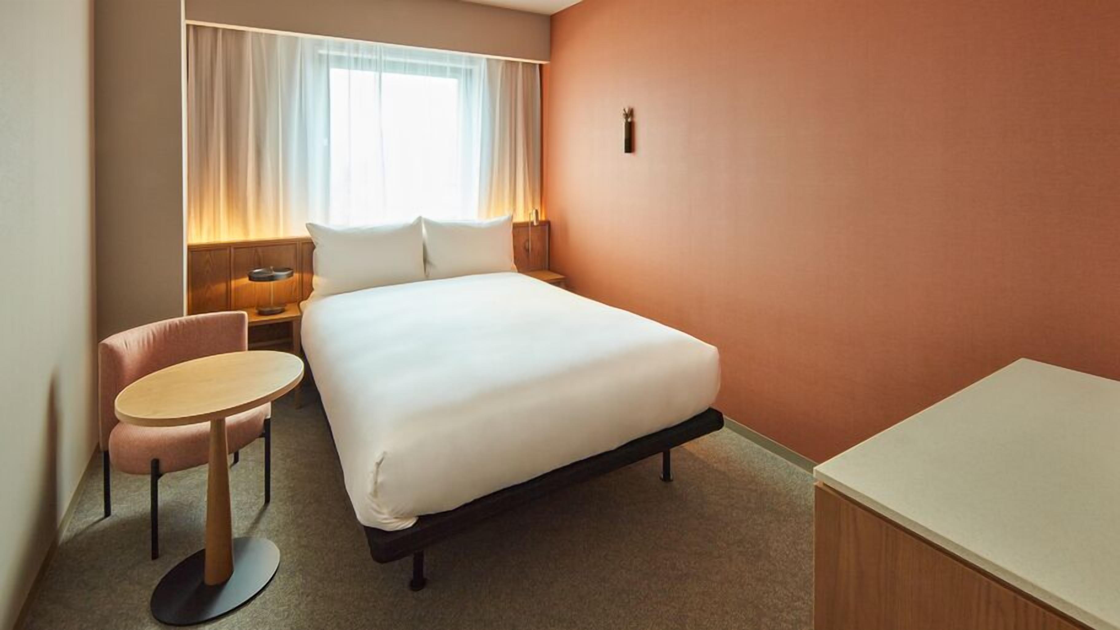 Une chambre d’hôtel comprenant un lit, un bureau, une chaise, une table de chevet et une fenêtre avec des rideaux.