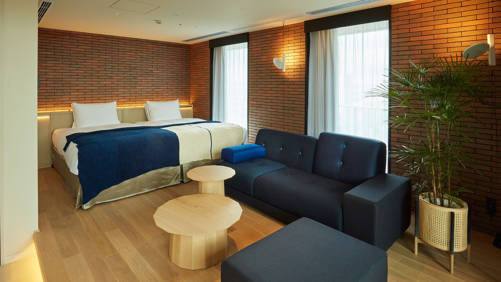 Une chambre d’hôtel moderne avec un mur en briques, un lit recouvert d’une couverture bleue, un canapé foncé, une table ronde en bois et une plante en pot.