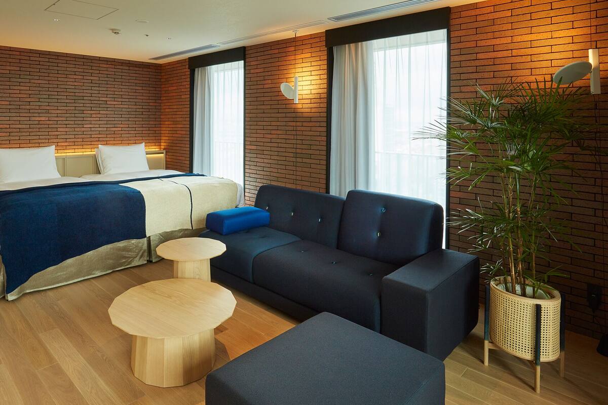Een moderne hotelkamer met een bakstenen muur, een bed met een blauwe deken, een donkere bank, een ronde houten tafel en een plant in pot.