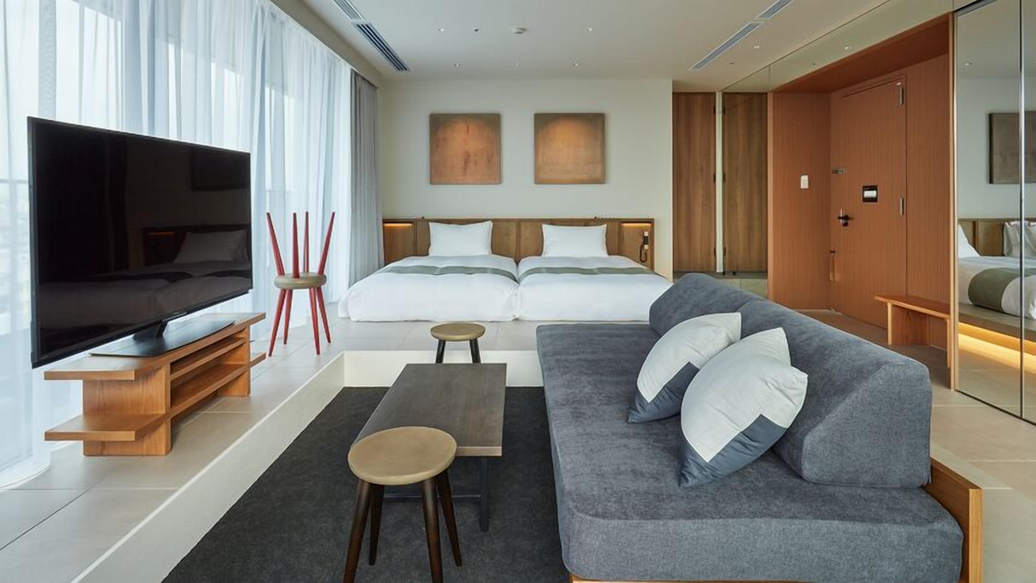 Une chambre d’hôtel moderne dotée d’un grand téléviseur à écran plat, d’un canapé gris avec des coussins blancs, d’une table basse en bois, d’une petite table ronde et d’un lit avec une literie blanche.