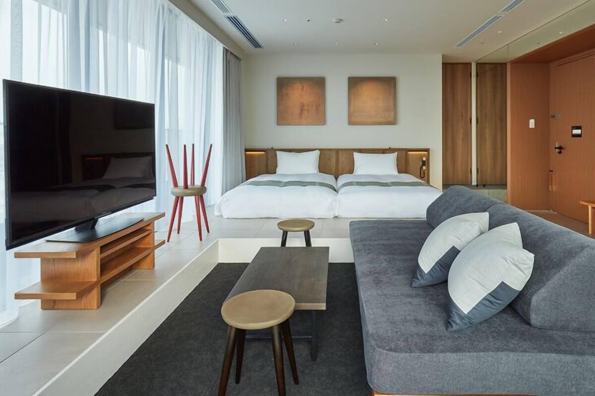 Een moderne hotelkamer met een grote flatscreen-tv, een grijze bank met witte kussens, een houten salontafel, een klein rond tafeltje en een bed met witte beddengoed.