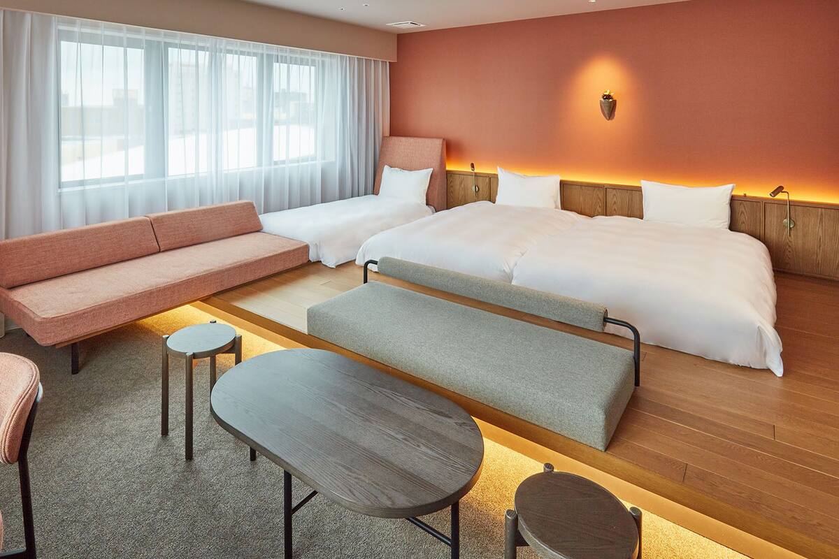 Een hotelkamer met een bank, twee eenpersoonsbedden, een kleine tafel, een bankje en een lamp.