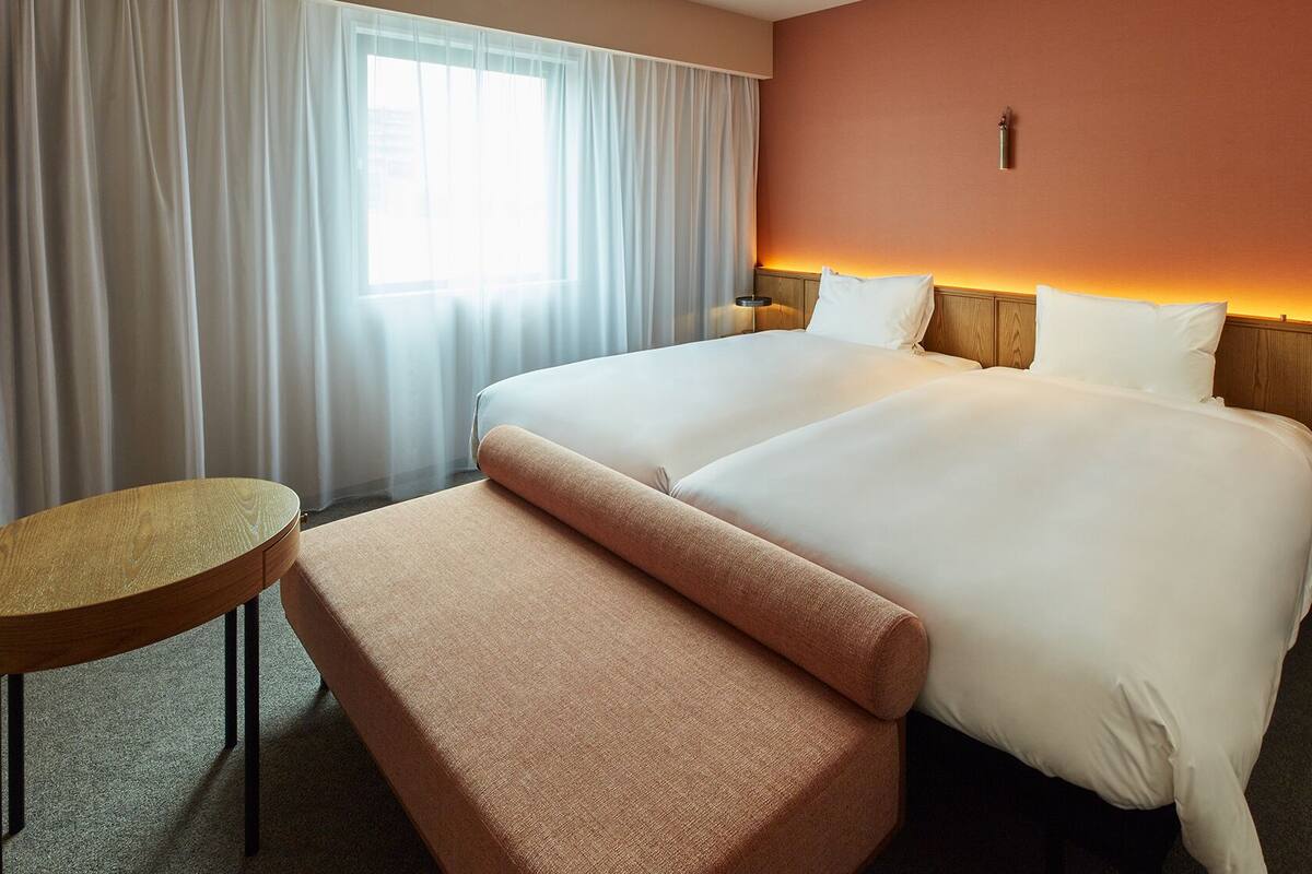 Een hotelkamer met een groot bed, een klein rond tafeltje, een bankje en een nachtkastje met een lamp.