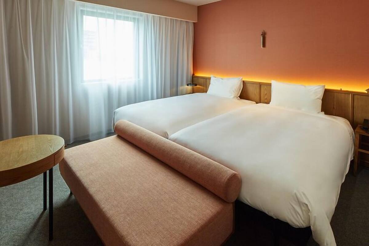 Een hotelkamer met een tweepersoonsbed, een klein rond tafeltje, een bankje en een nachtkastje met een lamp.