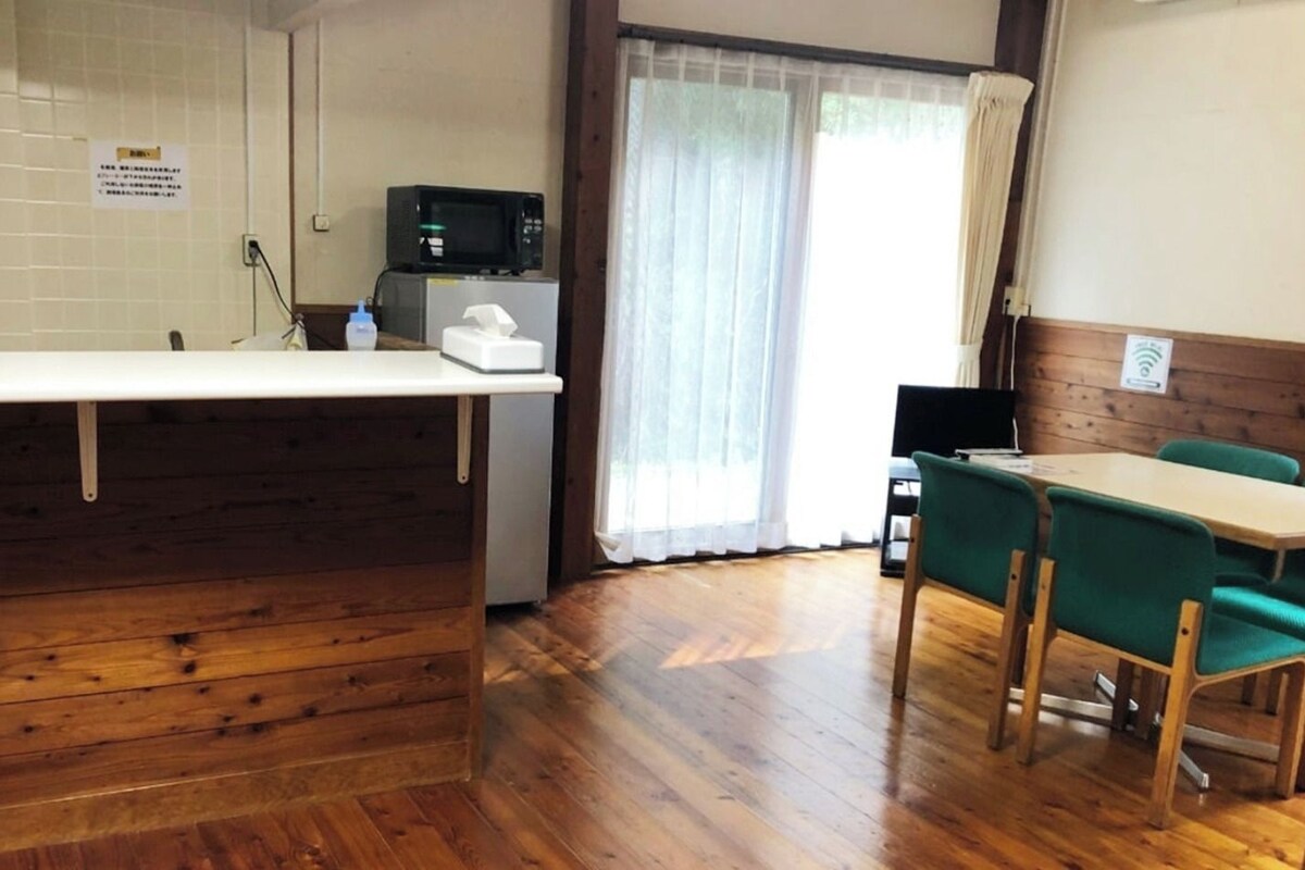 A wooden reception desk, a microwave, a refrigerator, and a dining area with green chairs.