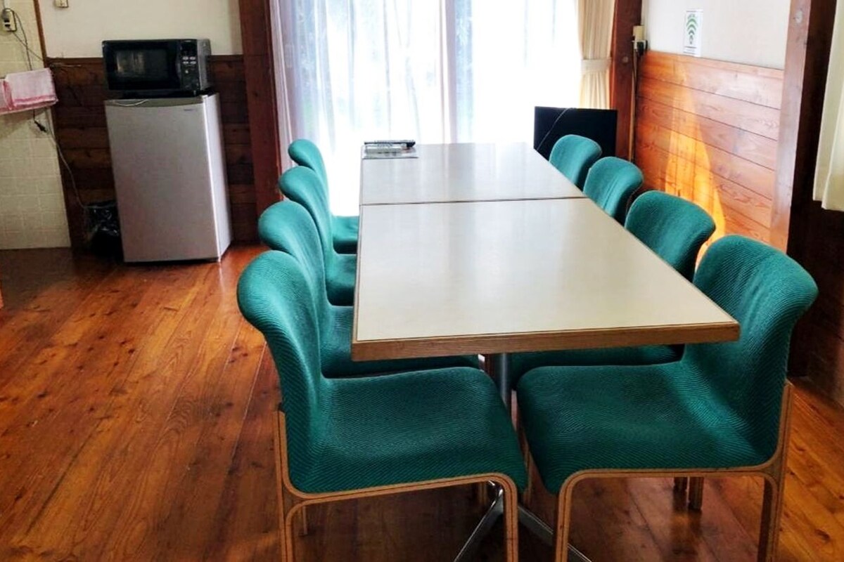 A dining area with a long table and six teal chairs, a small refrigerator, and a microwave on the left.