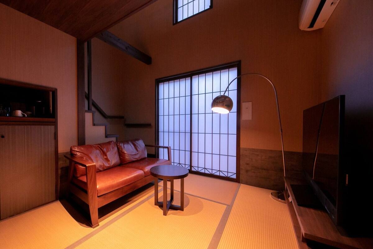 Ein Zimmer mit Tatami-Boden, einem Ledersofa, einem kleinen runden Tisch, einem Fernseher und einem Fenster mit Aussicht.