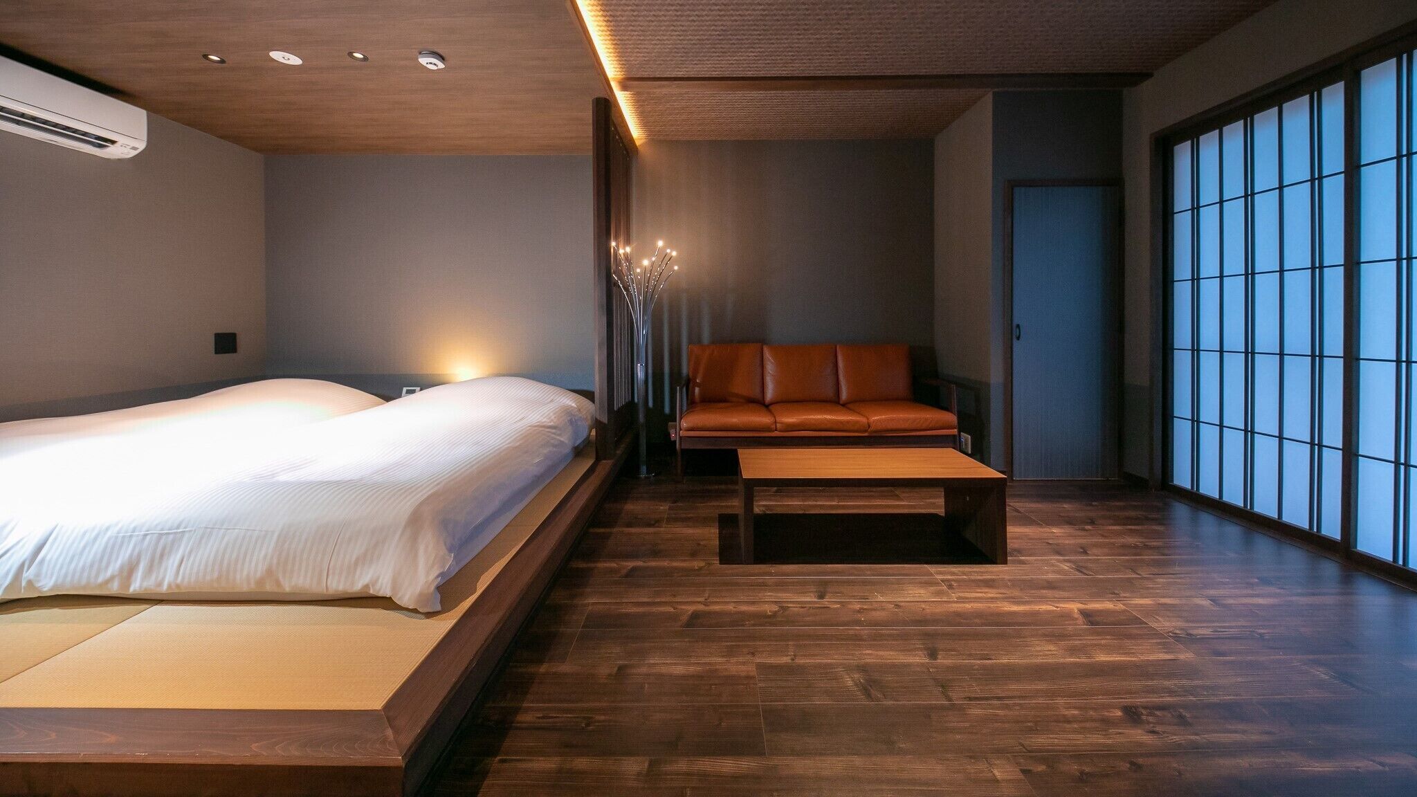 Moderni hotellihuone, jossa on suuri sänky, sohva, sohvapöytä ja näkymä ulos shoji-liukuoven läpi.