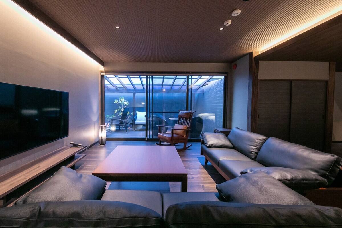 Ein modernes Wohnzimmer mit Flachbildfernseher, Ledersofa, Couchtisch aus Holz und einer Glastür zu einem Außenbereich.