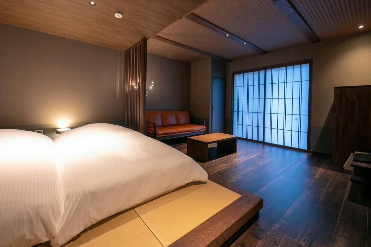 Ein modernes Hotelzimmer mit einem großen Bett, einer kleinen Sitzecke, Holzboden und einer Schiebetür aus Glas.