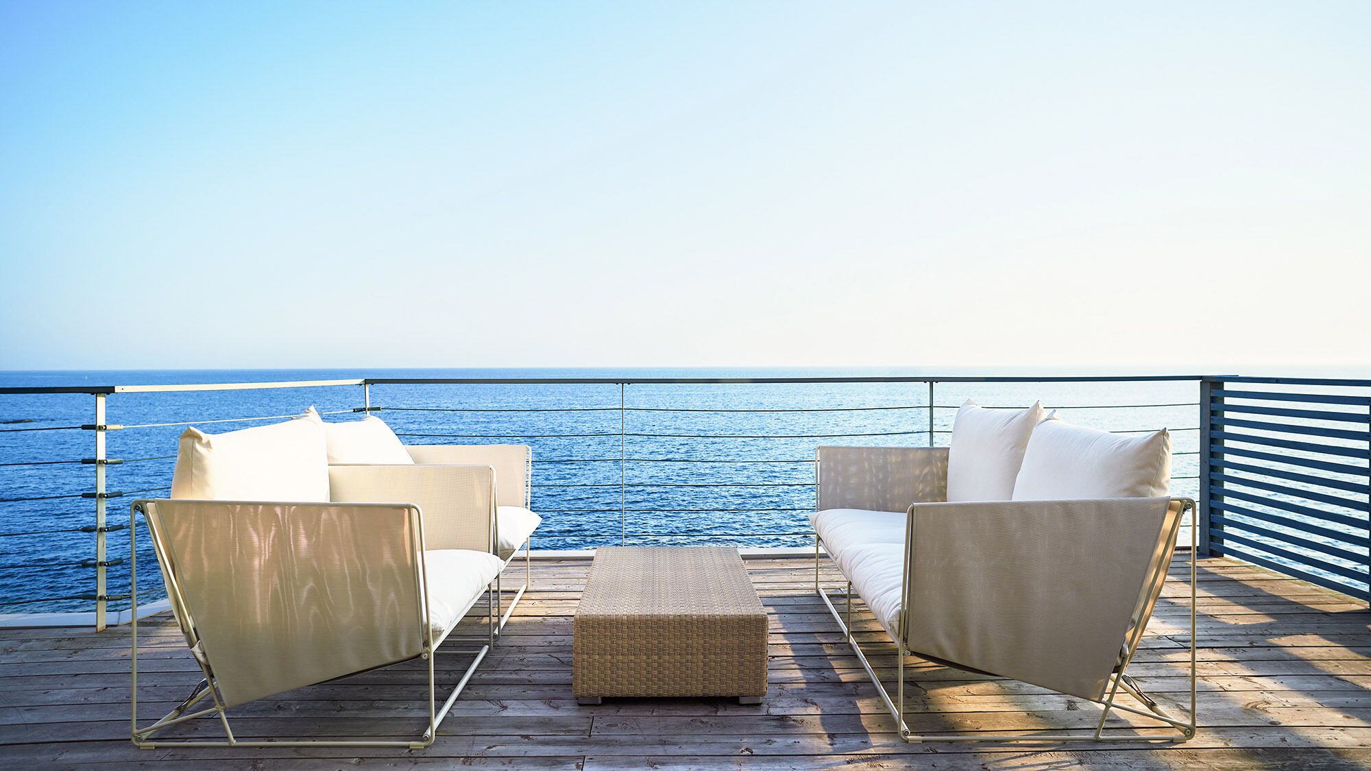 A deck with two beige chairs and a woven ottoman, overlooking a calm sea.