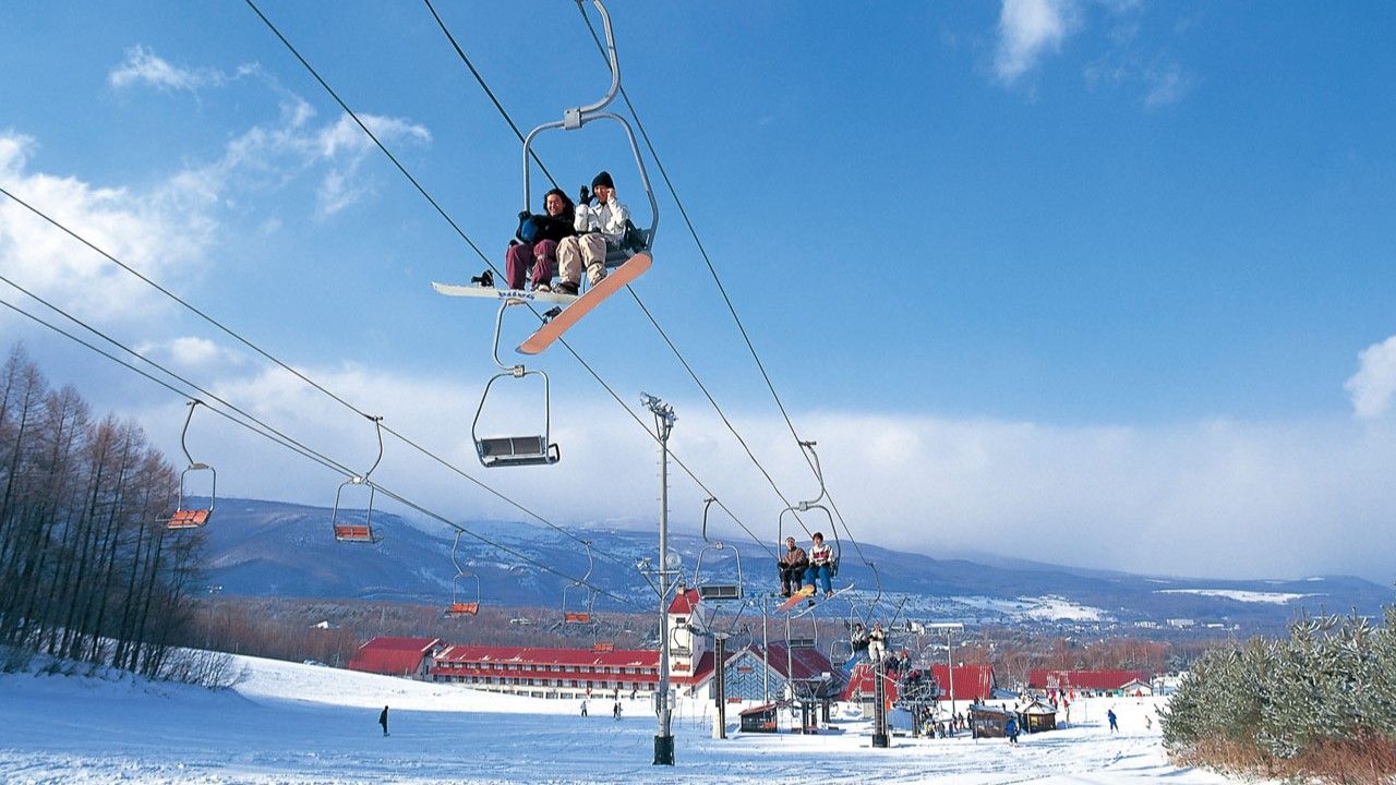 A ski lift with passengers, a snowy landscape, and buildings in the background.