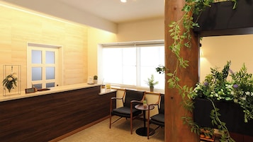 Un área de recepción con un mostrador de madera, una silla, un ventanal con persianas y una planta en maceta.