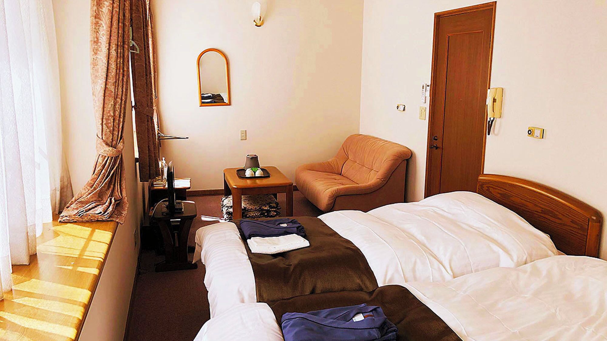 Habitación de hotel con dos camas, una silla, una mesita con lámpara, un espejo y una puerta.