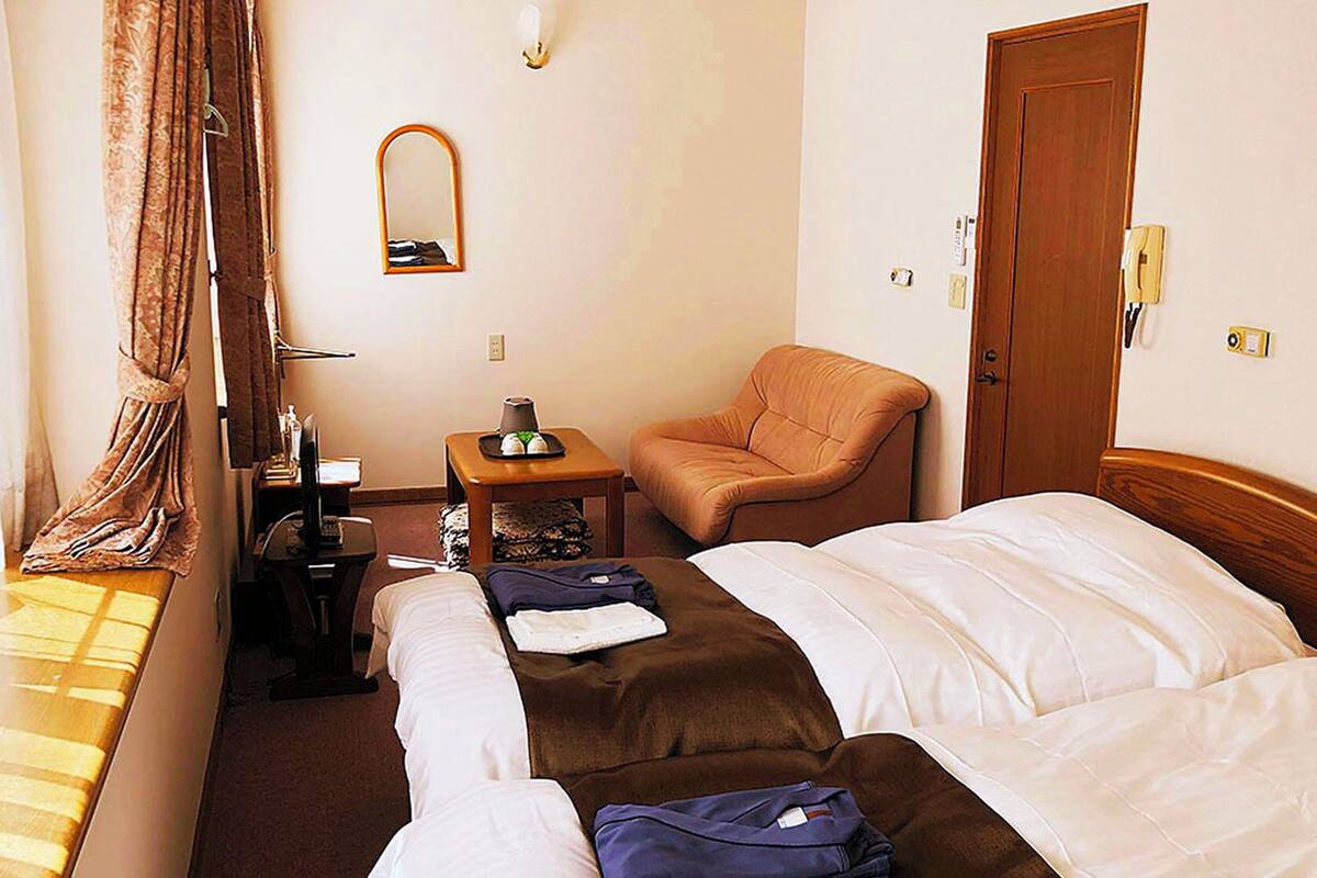 Een hotelkamer met twee bedden, een stoel, een kleine tafel met een lamp, een spiegel en een deur.