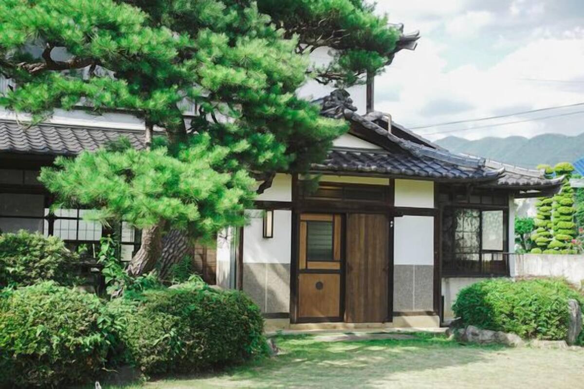 A traditional Japanese-style building with a wooden door, surrounded by lush greenery and a large tree.
