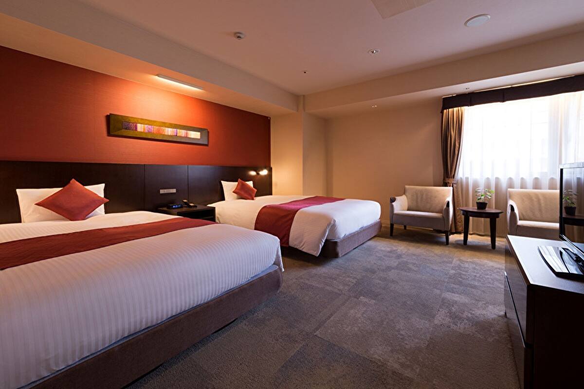 A hotel room with two beds, a TV, a desk, a chair, and a window with curtains.
