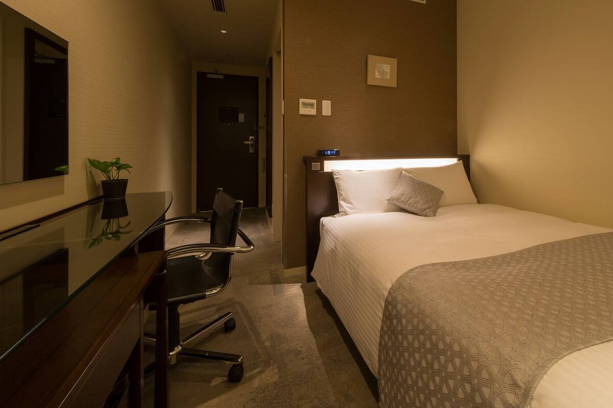 A hotel room with a bed, a desk, a chair, a mirror, and a wall-mounted clock.