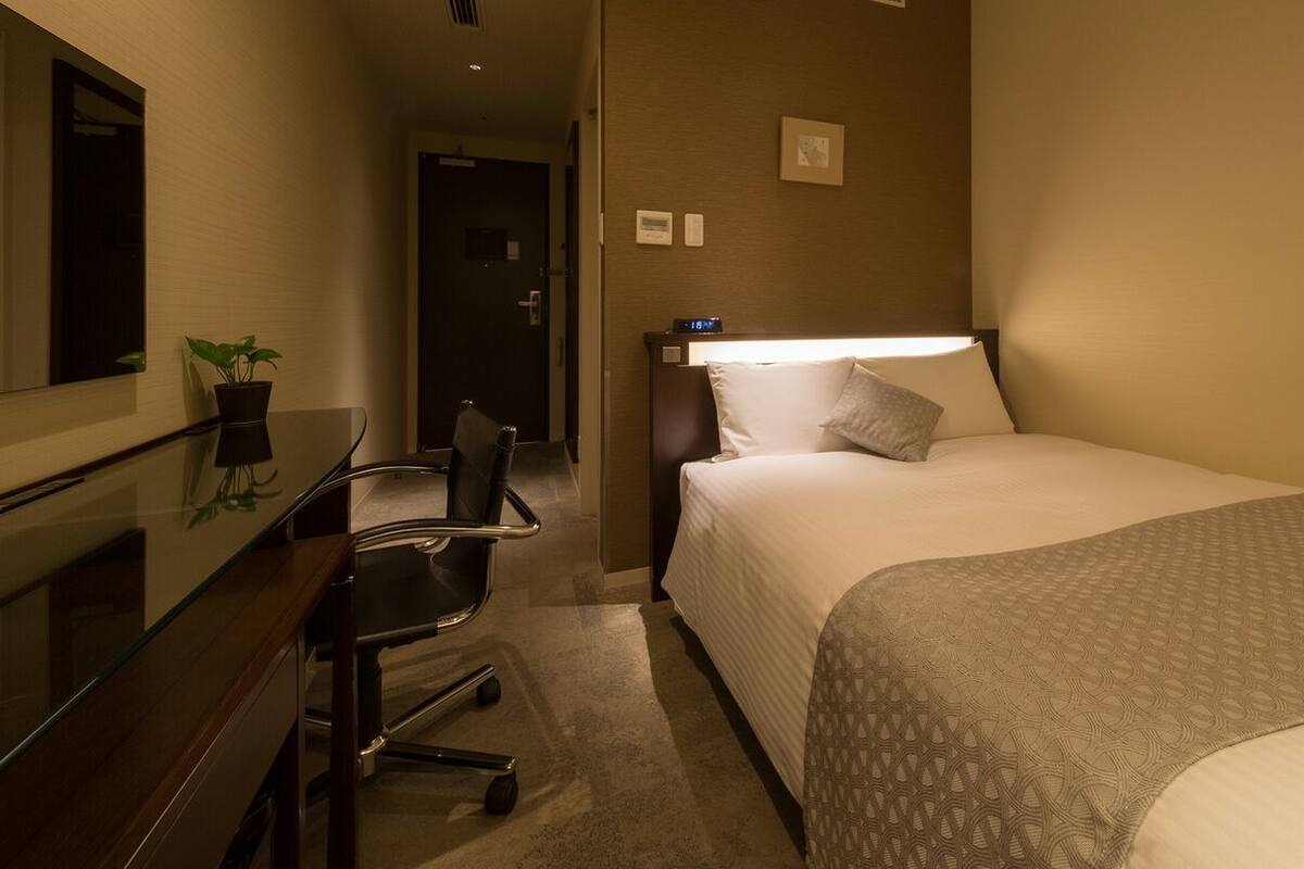 A hotel room with a bed, a desk, a chair, a potted plant, and a wall-mounted clock.