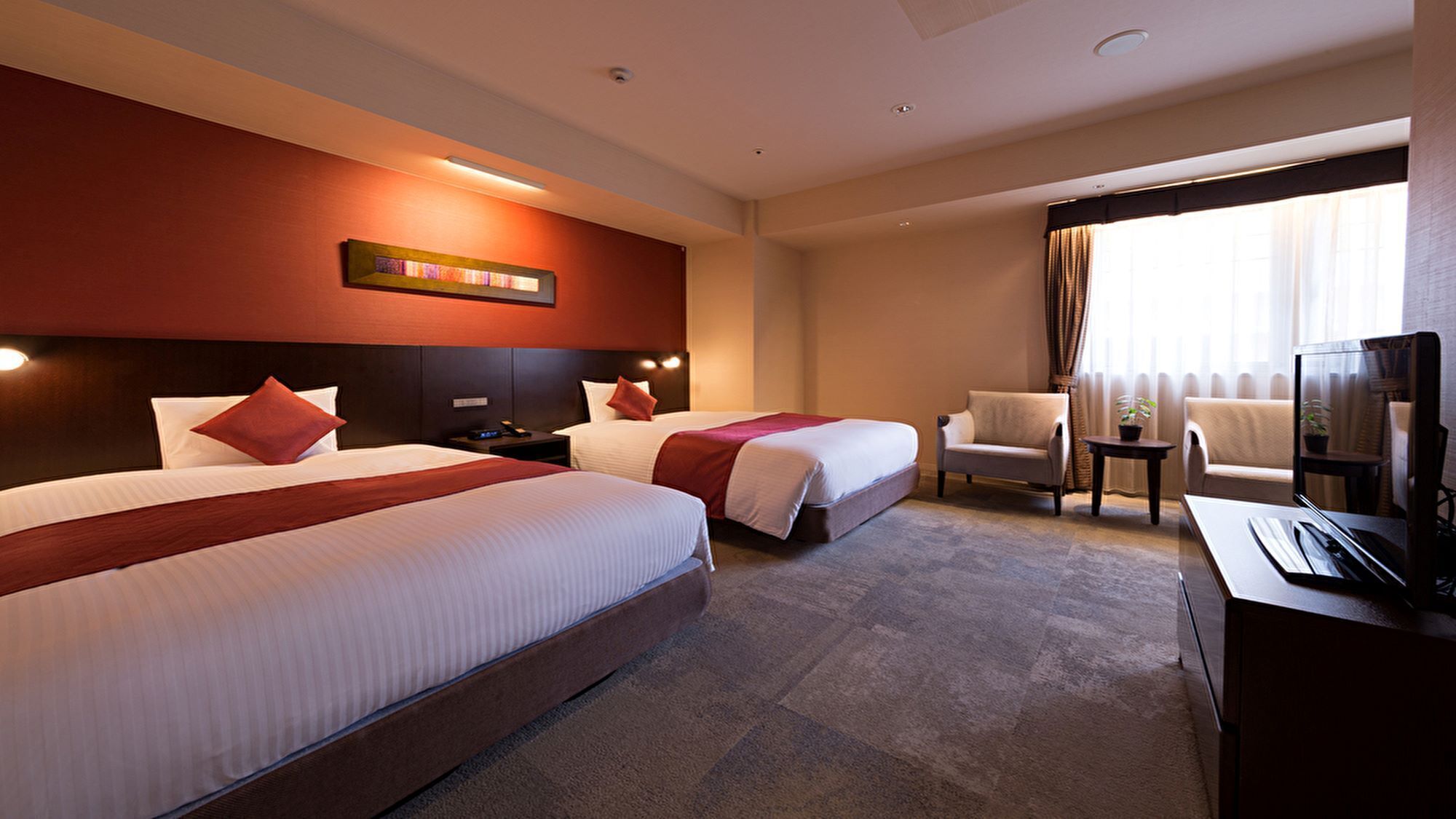 Ein Hotelzimmer mit zwei Betten, einem Schreibtisch mit Fernseher, einem Sessel und einem Fenster mit Vorhängen.