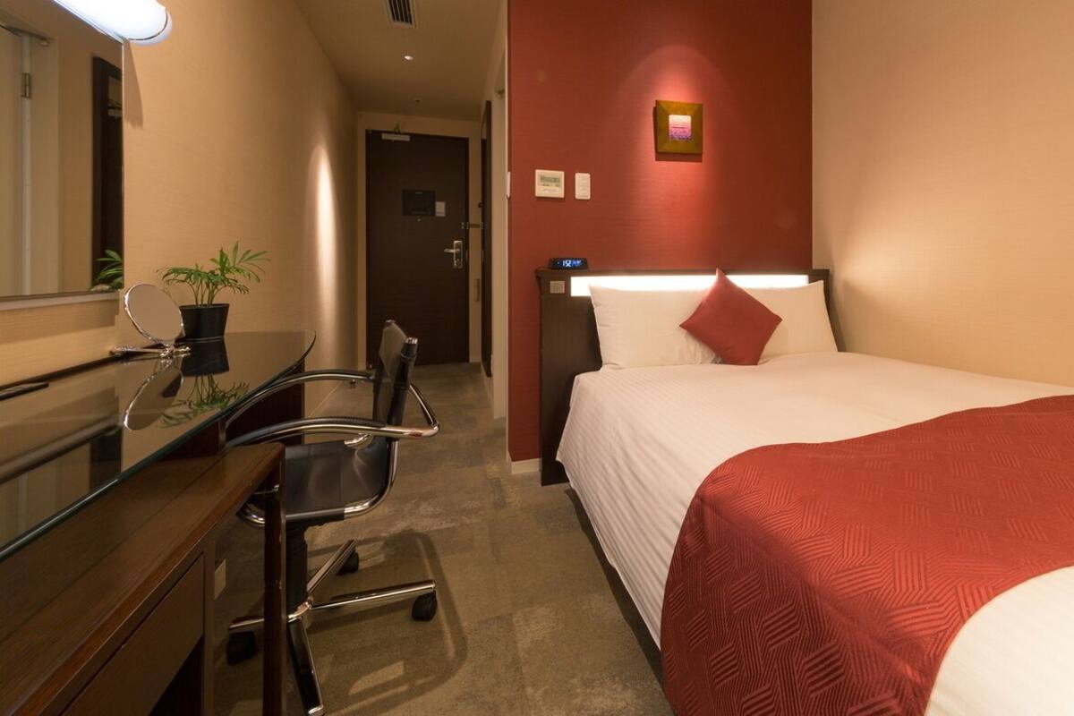 A hotel room with a bed, a desk with a chair, a mirror, a potted plant, and a nightstand.