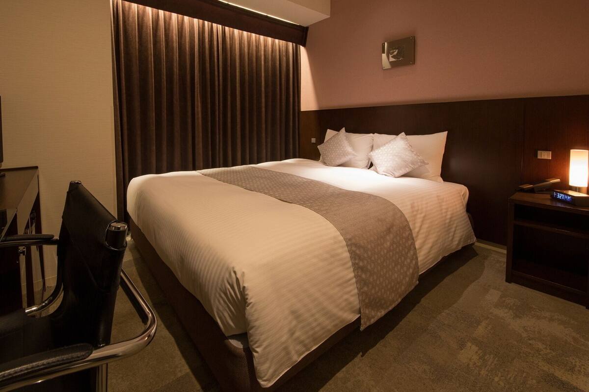 A hotel room with a bed, a desk with a chair, a television, a lamp, and a clock.