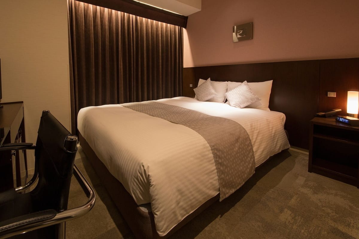 Habitación de hotel con cama, escritorio, silla, televisor, lámpara y reloj.