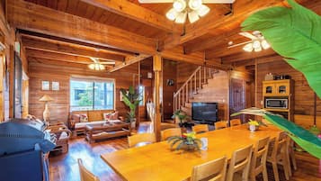 Interior de una cabaña de madera con comedor, asientos, televisor y escalera.