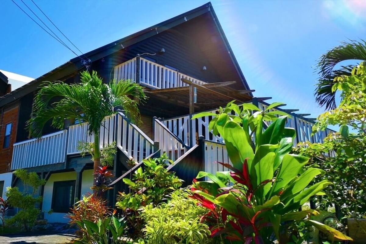 A two-story building with a balcony, surrounded by tropical plants.