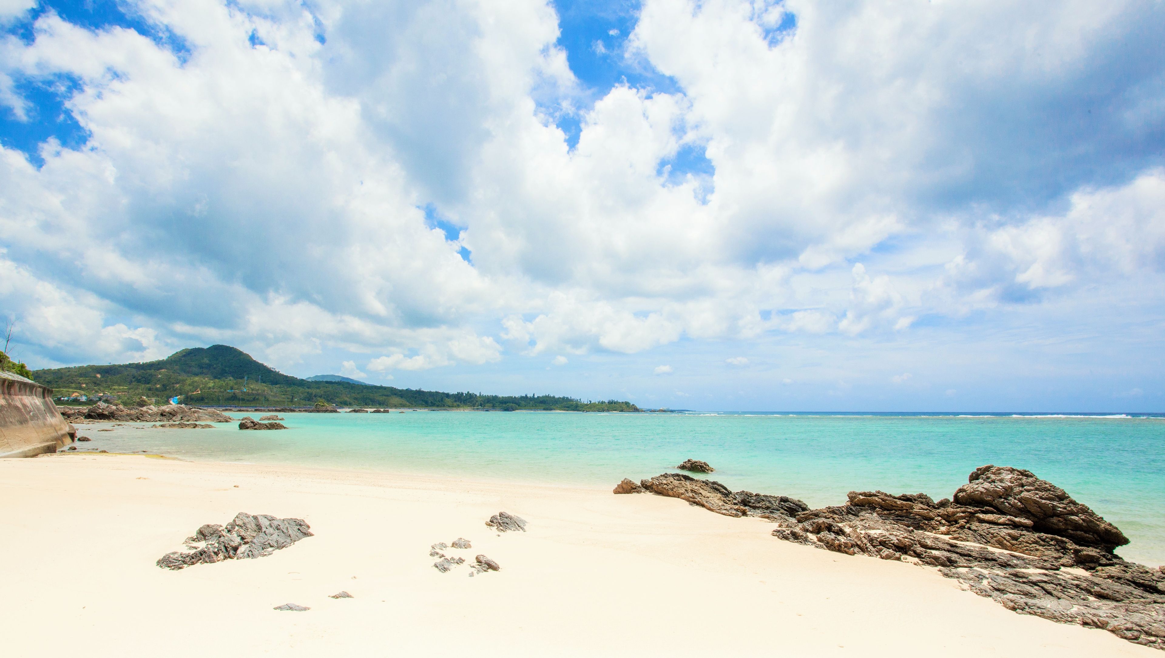 A beach with clear turquoise water, white sand, and scattered rocks.