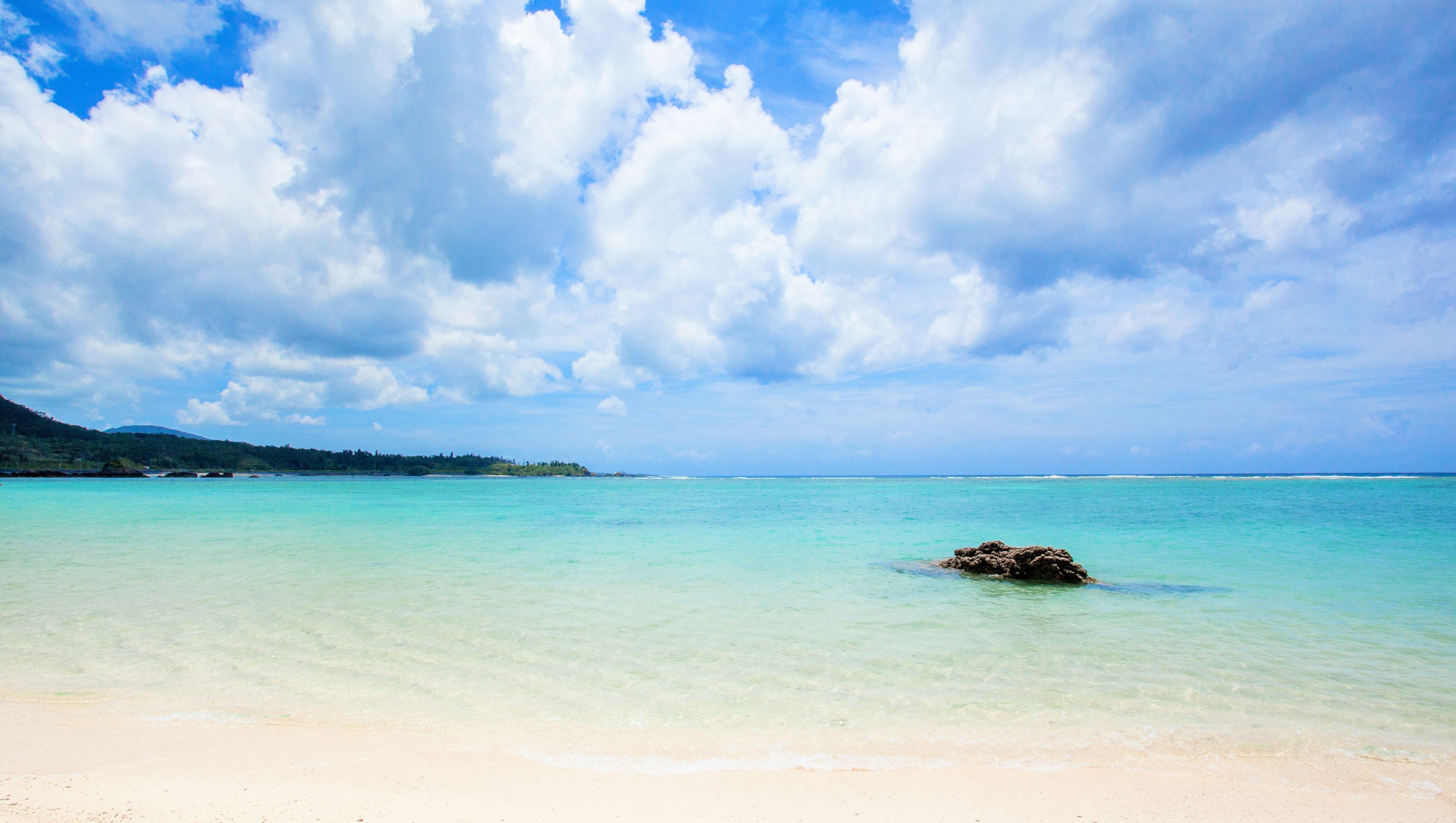 A beach with clear turquoise water, a sandy shore, and a rocky outcrop in the distance.