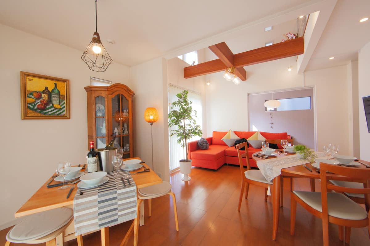 A dining area with a wooden table, chairs, and a dining set. There is a sofa and a potted plant in the living area.