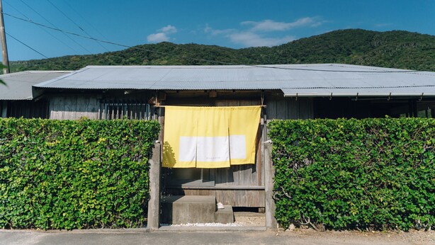 A building with a corrugated metal roof, a wooden door, and a yellow cloth hanging in front. - Denpaku Beach View Roof (Setouchi)