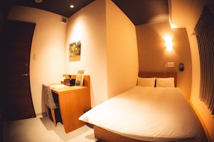 Una habitación de hotel con una cama, un escritorio, una silla, una mesita de noche, una lámpara de pared y un cuadro en la pared.