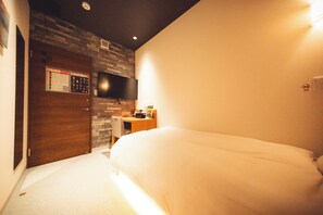 A hotel room with a bed, a desk, a flat-screen TV mounted on the wall, and a wooden door with a sign. - Baden Garden (Tokorozawa)