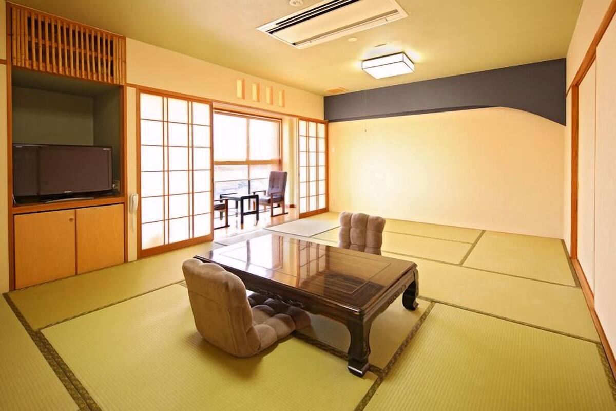 Una habitación japonesa tradicional con suelo de tatami, una mesa baja, sillas, un televisor y puertas correderas.