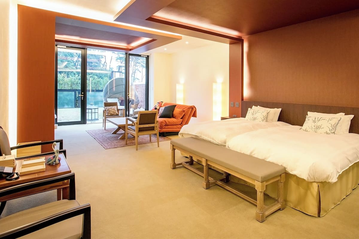 A hotel room with a large bed, a desk, a chair, a bench, and a balcony with outdoor seating.