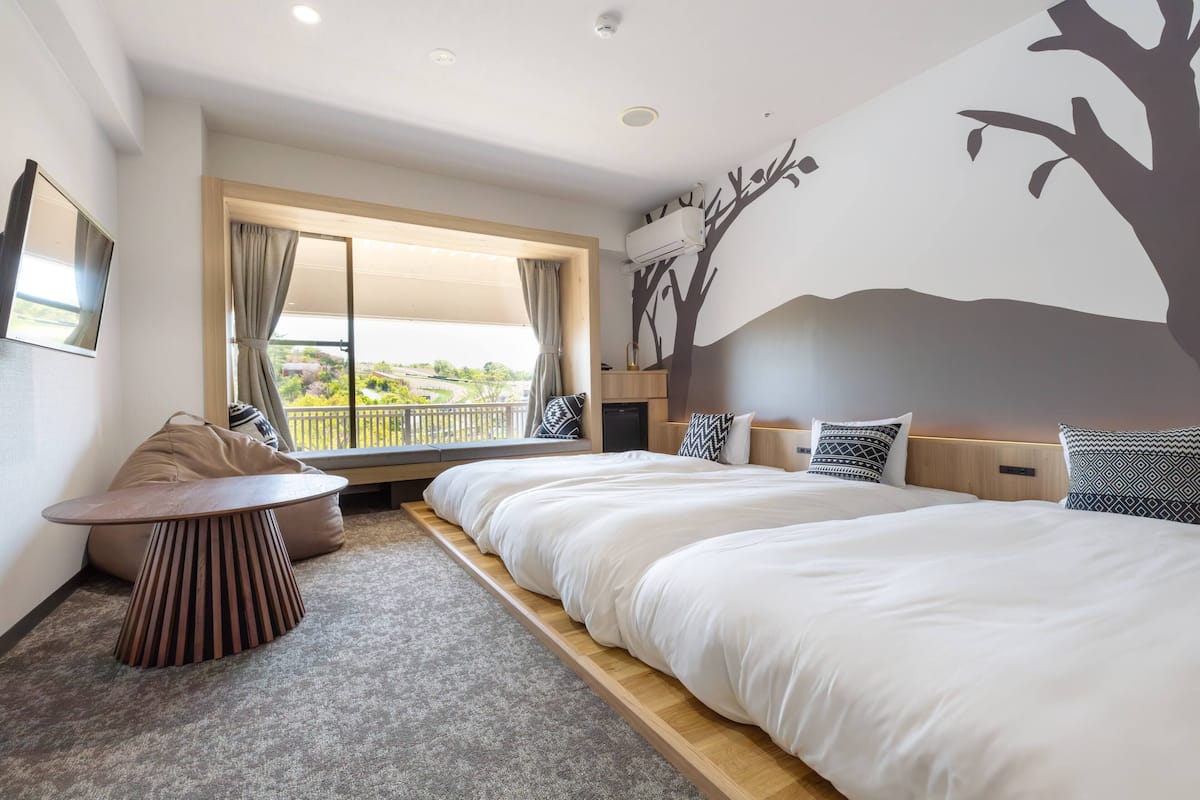 A hotel room with a large bed, a small table, a bean bag chair, a window with curtains, and a wall mural.