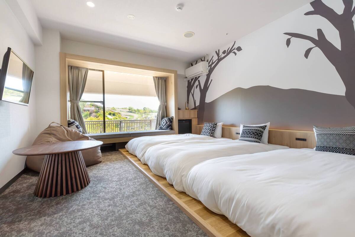 A hotel room with a large bed, a small table, a sofa, a window with curtains, and a wall mural.