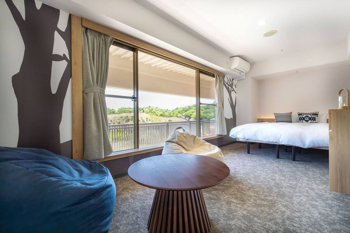 A bedroom with a bed, a small table, a bean bag, a balcony with a view, and a ceiling fan.