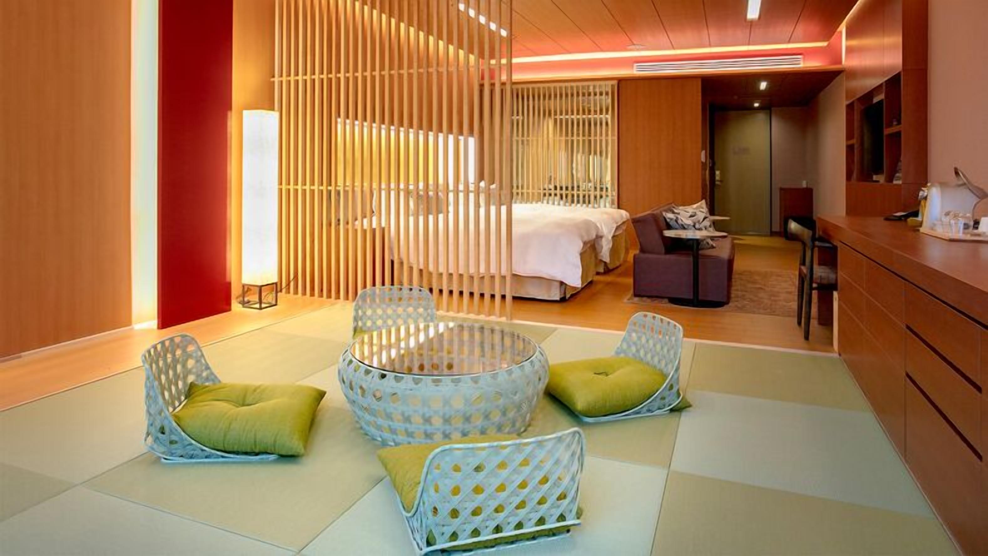 A hotel room with a bed, two seating areas with cushions, a desk, and a partition with vertical slats.