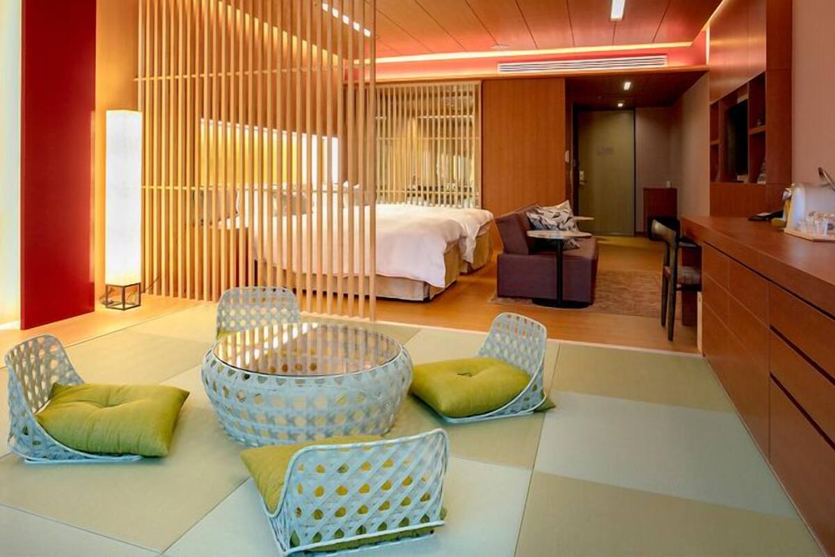 A hotel room with a bed, two seating areas with cushions, a desk, and a partition with vertical slats.