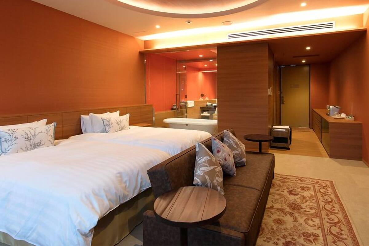A hotel room with a large bed, a sofa, a small round table, a bathroom with a bathtub, and a TV mounted on the wall.