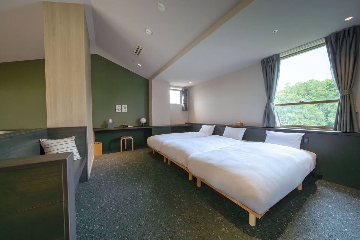 A hotel room with two beds, a small stool, a nightstand, a window with curtains, and a view of greenery outside.